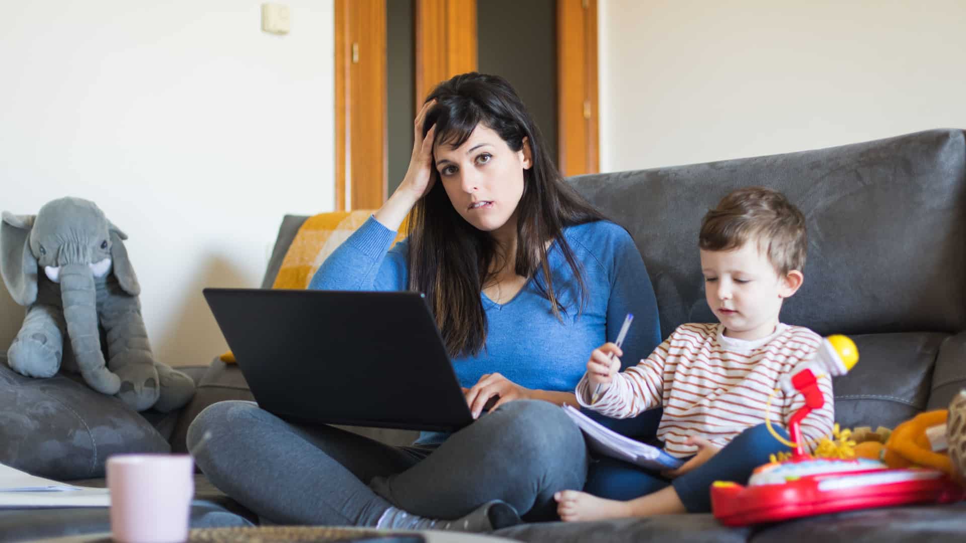 O mama coplesita sta pe canapea ei lucrand la un laptop, cu fiul ei tanar asezat langa ea.