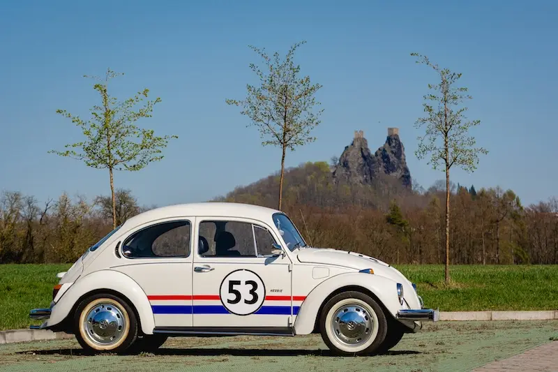 Vechiul Volkswagen Beetle alb, cu o dunga albastra si rosie si un numar de inceput 53 pe lateral.  Pe fundalul fotografiei, puteti vedea ruinele castelului Trosky din Paradisul Boem din Republica Ceha.
